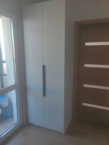 Изображение демонстрирует установленный шкаф в углу комнаты рядом с окном и дверью. Полка надежно закреплена, обеспечивая устойчивость и функциональность в интерьере.