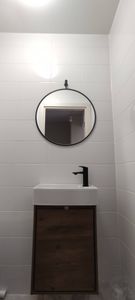 Круглое зеркало аккуратно закреплено на стене ванной комнаты над раковиной. Белая плитка создает светлый фон, подчеркивая стильный черный ободок.