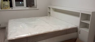 Собрана двуспальная кровать с белым изголовьем и встроенными полками. Все элементы надежно закреплены, матрас установлен, мебель готова к использованию.