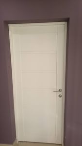 Установлена новая белая дверь, закреплены петли, врезан замок, ручка прикреплена. Проем оформлен наличниками, все элементы подогнаны, конструкция функционирует плавно и надежно.