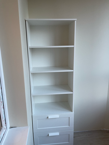 Собран белый стеллаж с четырьмя полками и двумя выдвижными ящиками, установлен в углу комнаты. Конструкция устойчива, детали плотно подогнаны, обеспечена надежность и функциональность.