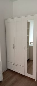 Собран белый шкаф с тремя дверцами и зеркалом, установлен на деревянный пол. Все элементы надежно закреплены, ручки прикручены, дверцы ровно висят, конструкция устойчива.