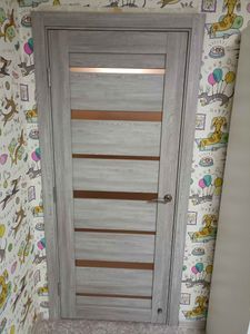 Установлена новая межкомнатная дверь с петлями, замком и ручкой. Полотно ровно закреплено в проеме, обеспечивая плавное открывание и закрывание. Монтаж завершен, все элементы на месте.