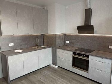 Изображение современной кухни с серыми шкафами и встроенной техникой. На столешнице раковина, плита, вытяжка. Интерьер выполнен в минималистичном стиле с акцентом на функциональность.