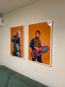 Изображены две яркие картины в белых рамках, аккуратно закрепленные на светлой стене над зеленым диваном.