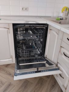Изображение показывает кухню с открытой посудомоечной машиной, встроенной в мебель. Видны деревянный пол и белая столешница. На заднем плане розетки и кухонные принадлежности.