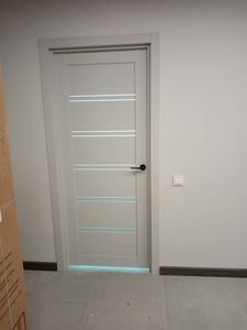 Установлена новая дверь с ручкой, петли закреплены, коробка выровнена, наличники смонтированы, замок функционирует, полотно ровно висит, щели отсутствуют, конструкция завершена.