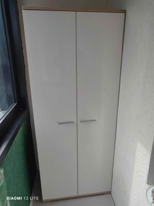 Изображение показывает установленный шкаф на балконе. Белые двери с ручками, аккуратно размещены у стены. Рядом окно, свет падает на поверхность.