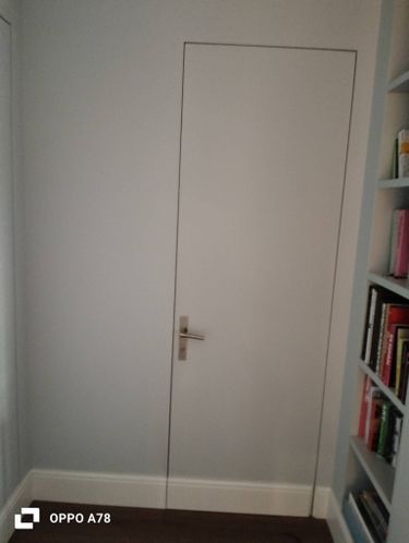 Установлена скрытая дверь, гармонично вписывающаяся в стену. Дверное полотно окрашено в тон, ручка металлическая. Рядом книжная полка, создающая уютную атмосферу.