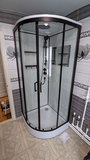 Установлена современная душевая конструкция с прозрачными стенками, черными рамами и удобными дверцами. Поддон белый, аккуратно размещен на полу. Стены облицованы плиткой с узором.