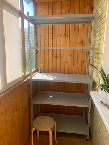 Собран металлический стеллаж с четырьмя полками на застеклённой лоджии. Конструкция установлена в углу, обеспечивая устойчивость и функциональность для хранения.