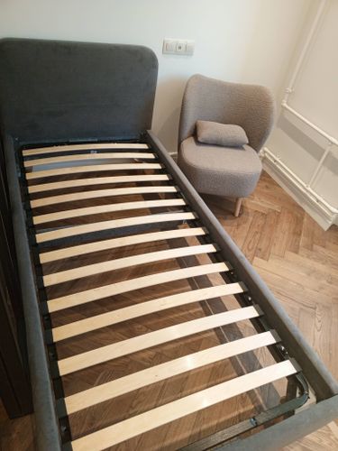Собрана кровать с деревянными ламелями, установлена в комнате. Рядом стоит мягкое кресло. Пол покрыт паркетом. Стены светлые, розетки на месте. Всё выглядит аккуратно и завершено.