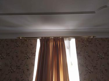 Закреплен металлический держатель для штор на стене. Установлены крепления, обеспечивающие надежность конструкции. Гардины висят ровно, создавая уютную атмосферу в комнате.