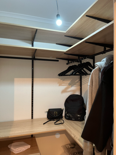 В комнате закреплены металлические направляющие для полок, установлены деревянные полки и перекладины для вешалок. Пространство организовано для хранения одежды и аксессуаров.