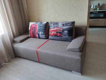 Собран диван: установлены каркас, подлокотники, подушки. Проверена устойчивость, закреплены все детали. Готов к использованию, стоит в комнате у стены, рядом окно с занавесками.