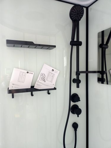 Изображение душевой кабины с черной лейкой и полками. На полках лежат инструкции. Стены белые, гладкие. Установлены смесители и держатель для душа.