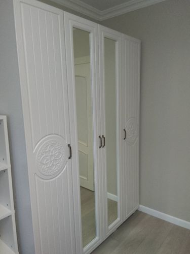 Собран белый шкаф с зеркальными дверцами и декоративными элементами, установлен в углу комнаты. Ручки прикреплены, конструкция устойчива, готова к использованию.