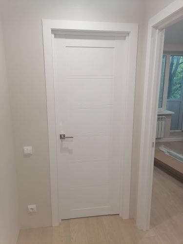 Установлена новая белая дверь с ручкой, закреплена в проеме. Осуществлена подгонка полотна, монтаж коробки и наличников. Все элементы выровнены и зафиксированы, обеспечена плавная работа.