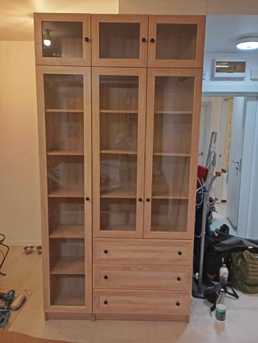Собран высокий шкаф с полками и стеклянными дверцами, установлены ящики, закреплены ручки. Все детали надежно соединены, конструкция устойчива и готова к использованию.