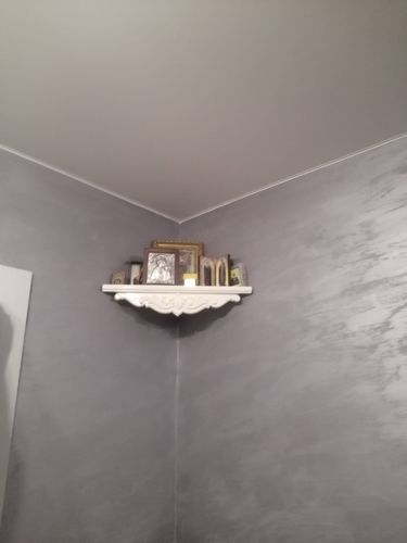 Угловая полочка закреплена на стене, украшена рамками и книгами. Стены серые, создают контраст с белым элементом. Работа завершена аккуратно, предметы расположены устойчиво.