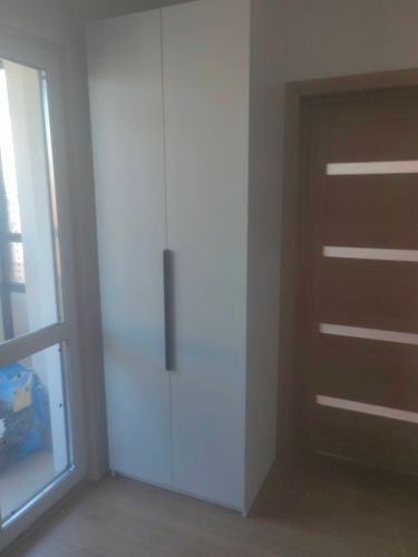 Изображение демонстрирует установленный шкаф в углу комнаты рядом с окном и дверью. Полка надежно закреплена, обеспечивая устойчивость и функциональность в интерьере.