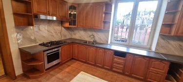 Изображена кухня с деревянными шкафами, газовой плитой, вытяжкой, мойкой и окном с решеткой. Столешница темная, фартук из светлого камня. Пол выложен плиткой.