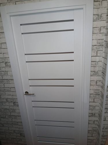 Установлена белая дверь с горизонтальными полосами, вмонтирована ручка, закреплён наличник, обеспечена ровная посадка в проём, завершён монтаж на кирпичной стене.