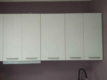 Установлены навесные шкафы с белыми фасадами и металлическими ручками, закреплены на стене. Смонтирован смеситель, завершена сборка кухонного гарнитура.