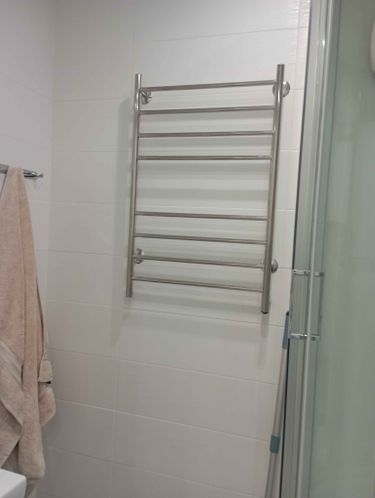 На стене ванной комнаты закреплен металлический полотенцесушитель, подключенный к электросети. Устройство надежно установлено и готово к использованию.