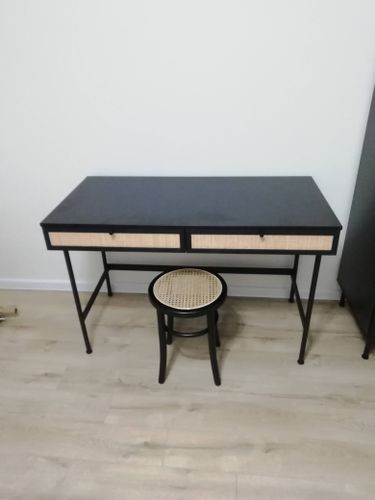 Собран стильный черный стол с двумя ящиками, дополненный круглым табуретом с плетеным сиденьем, установлен на светлом ламинате у белой стены.