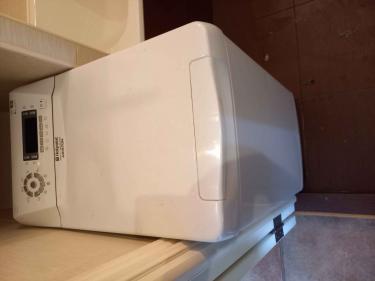 На изображении показана стиральная машина Hotpoint Ariston, установленная в углу ванной комнаты. Машина белого цвета, с верхней панелью управления и цифровым дисплеем.