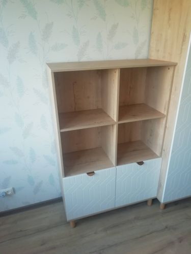Собран деревянный стеллаж с четырьмя полками и двумя нижними закрытыми секциями. Установлены ножки, дверцы закреплены. Готовая конструкция стоит на полу, прилегает к стене.