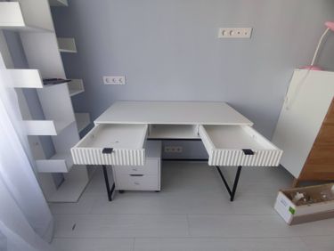 Собран белый стеллаж с несколькими полками, установлен рядом с письменным столом. Все элементы надежно закреплены, обеспечивая устойчивость и функциональность.