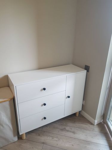 Собран белый комод с тремя ящиками и дверцей. Установлены ручки, закреплены ножки. Мебель размещена в углу на деревянном полу, рядом окно. Работа завершена аккуратно.