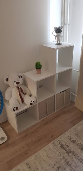 Собран белый модульный стеллаж с несколькими секциями, установлены декоративные элементы: мягкая игрушка, растение в горшке и фигурка. Компактно размещён на полу у стены.