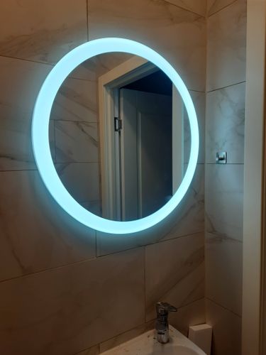 На стене ванной комнаты закреплено круглое зеркало с подсветкой. Установлено ровно, надежно, без видимых креплений. Стильный элемент интерьера завершает облик помещения.