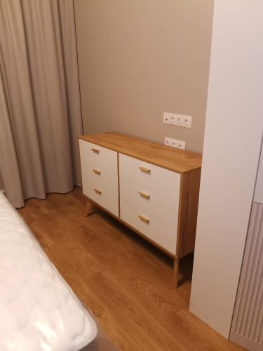 Собран деревянный шкаф с шестью ящиками, установлен в спальне. Все детали соединены, ручки прикреплены, конструкция устойчива, поверхность ровная, размещен рядом с кроватью и розетками.