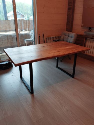 Изготовлен и установлен новый деревянный стол с металлическими ножками в светлой комнате. Пол из ламината, вид из окна на террасу и лес. Мебель гармонично вписана в интерьер.