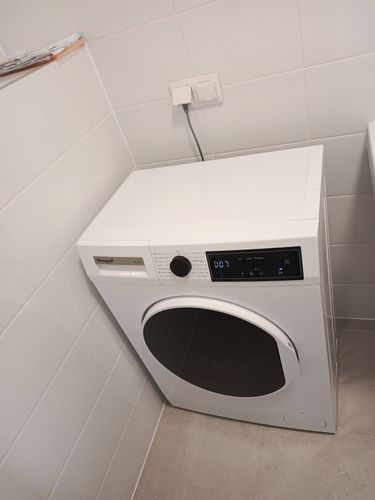 Изображена стиральная машина, подключенная к электросети и водопроводу. Устройство установлено на плиточном полу в углу помещения с белыми стенами.