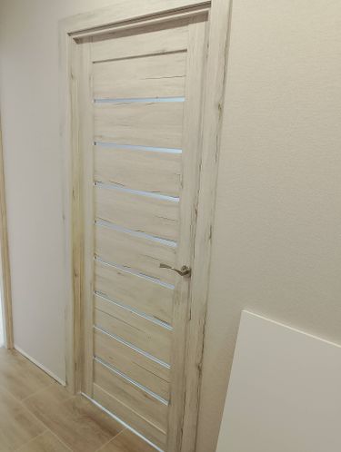 Установлена светлая межкомнатная дверь с горизонтальными вставками и металлической ручкой. Полотно аккуратно вмонтировано в проем, обеспечивая плавное открывание и закрывание.