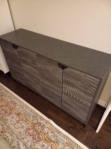 Собран новый предмет мебели: установлены панели, закреплены дверцы, прикручены ручки, выровнены ножки. Готовое изделие стоит на ковре, гармонируя с интерьером.