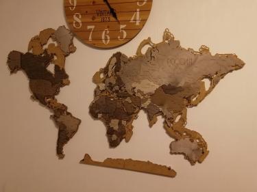 На стене закреплена деревянная карта мира, состоящая из нескольких частей. Элементы размещены ровно, создавая гармоничную композицию. Карта дополнена стильными часами.