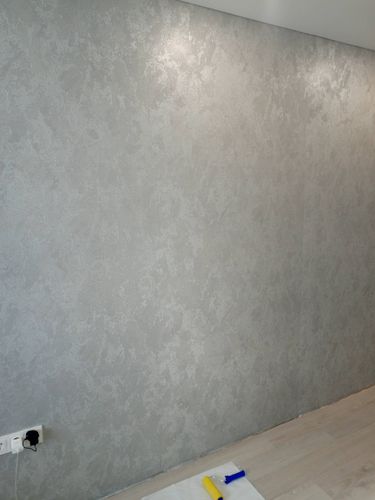 На стене аккуратно наклеены серые обои с текстурным узором. Поверхность ровная, без видимых швов и пузырей. Работа завершена, инструменты убраны, пол чистый.