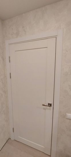 Установлена белая межкомнатная дверь с одной створкой, закреплены петли, ручка, наличники. Все элементы подогнаны, конструкция ровно размещена в проеме, обеспечивая плавное открывание.