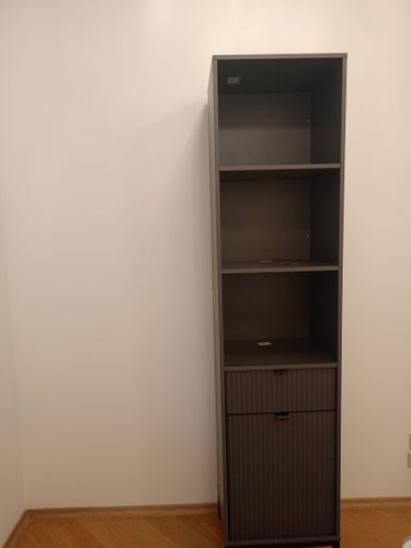 Собран высокий шкаф с полками и выдвижным ящиком, установлен в углу комнаты. Все детали надежно закреплены, конструкция устойчивая, готова к использованию.