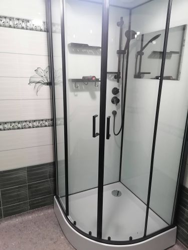 Установлена новая душевая кабина с прозрачными стенками и черными рамами. Поддон заменен, смеситель и лейка закреплены. Пол и стены облицованы плиткой, добавлены полки для удобства.