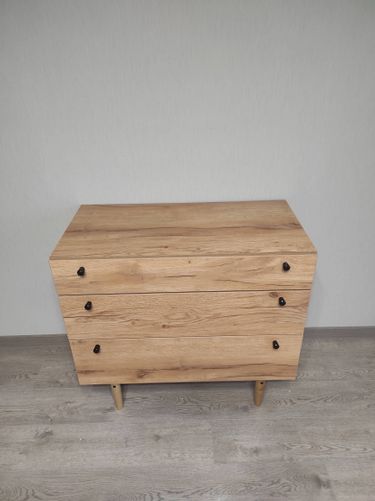 Собран деревянный комод с тремя ящиками, установленными ручками и устойчивыми ножками. Все детали соединены, конструкция выровнена и проверена на устойчивость.