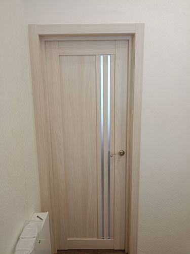 Установлена светлая дверь с вертикальными вставками, закреплена ручка, отрегулированы петли, смонтирован наличник. Работа завершена, конструкция надежно зафиксирована в проеме.