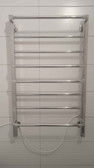 На стене ванной закреплен металлический прибор для сушки полотенец, подключенный к электросети. Устройство надежно установлено, кабель аккуратно проложен.