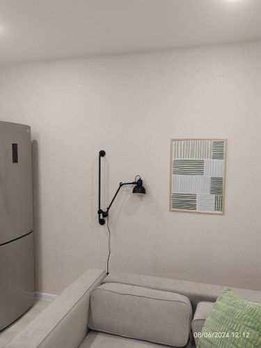 На стене закреплена настенная лампа с подвижным кронштейном, обеспечивающая освещение над диваном. Рядом висит картина в светлой рамке, создавая уютную атмосферу в комнате.